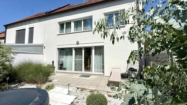 Hausfassade mit großer Terrasse, Whirlpool und Gartenbereich unter blauem Himmel.