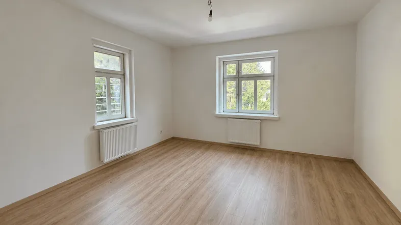 Helles, leeres Zimmer mit Laminatboden und zwei Fenstern, bereit zur individuellen Gestaltung.