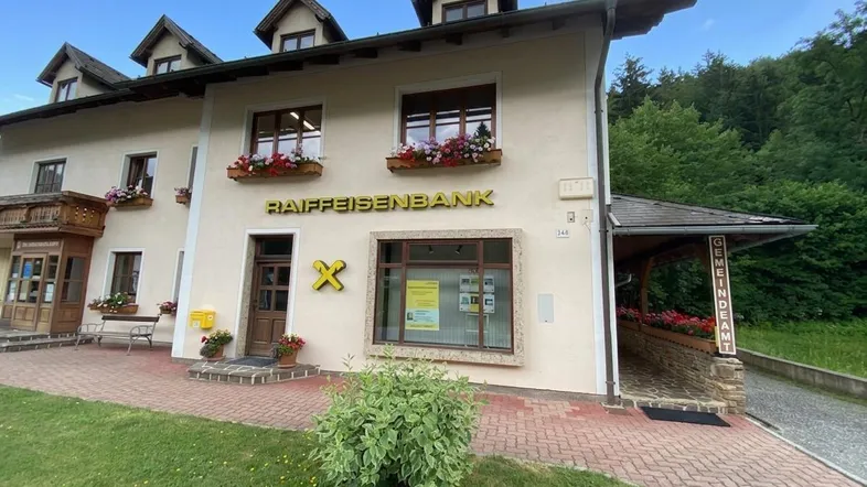 Das Gebäude der Raiffeisenbank zeigt traditionelle Architektur mit Blumenkästen und ist von viel Grün umgeben.