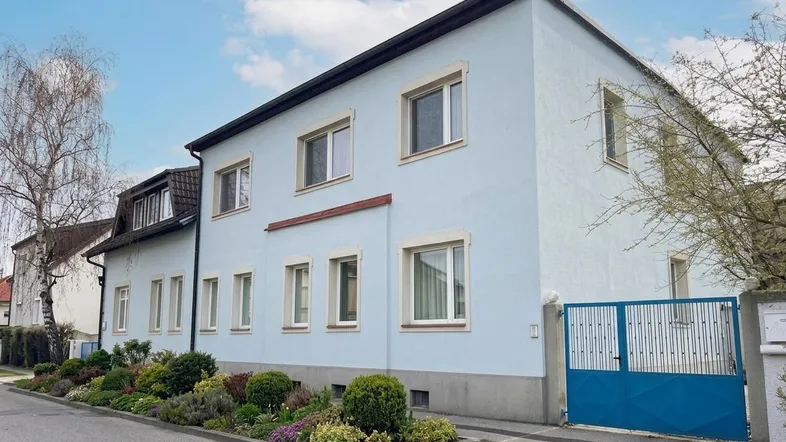 Ansicht des hellblauen Gebäudes von der Straße mit gepflegtem Vorgarten und blauem Tor.
