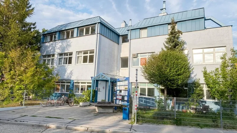 Außenansicht eines mehrstöckigen Gebäudes mit blauen Dachelementen und umgebender Vegetation.