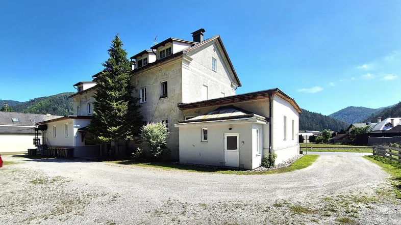 Großes Wohngebäude mit heller Fassade und Satteldach, umgeben von Bergen und Natur.