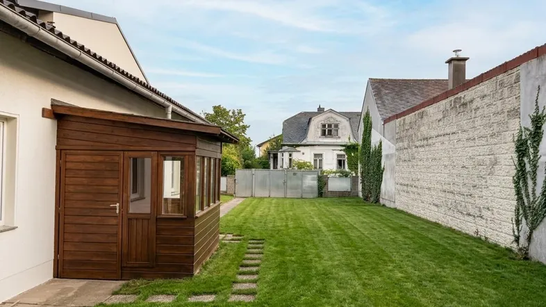 Gepflegter Gartenbereich mit Rasenfläche, Holzschuppen und Steinfliesenweg zwischen den Gebäuden.