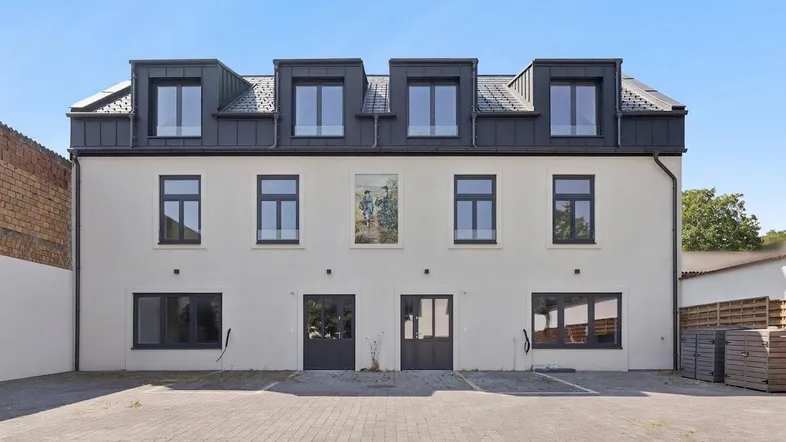 Moderne Fassade eines neuwertigen Gebäudes mit weißem Putz, dunklen Fenstern und Mansarddach.
