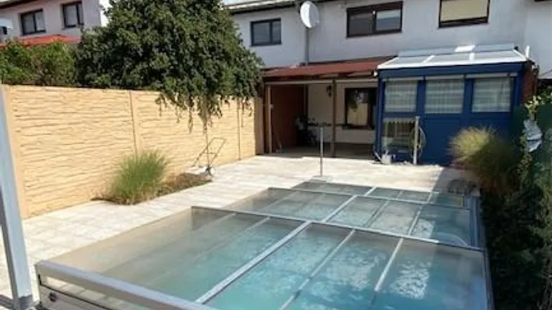 Hinterhof mit überdachtem Pool und Holzterrasse unter strahlend blauem Himmel.
