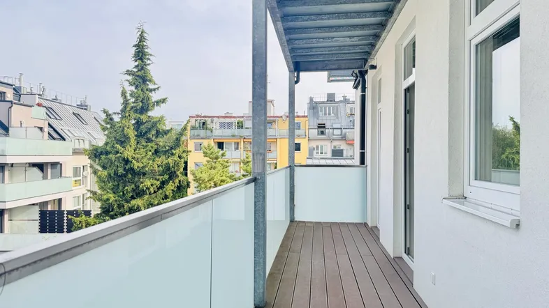 Geräumiger Balkon mit Holzboden und Glasgeländer bietet einen Blick auf die umliegenden Gebäude und Bäume.
