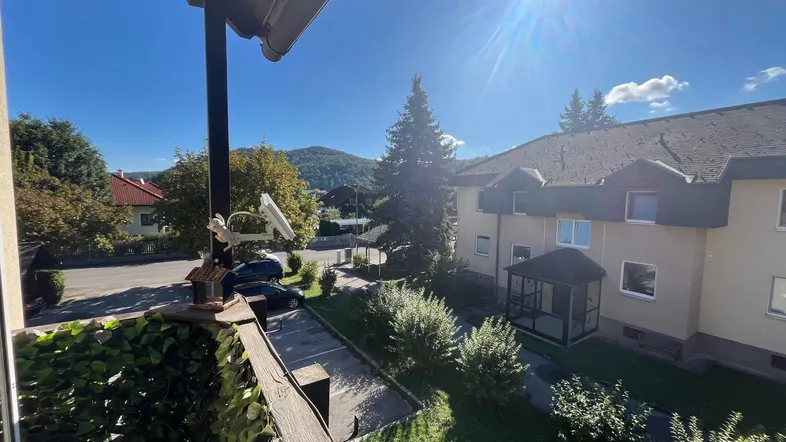 Sonniger Ausblick vom Balkon auf die Umgebung mit Parkplätzen, weiteren Gebäuden und grünen Hügeln.