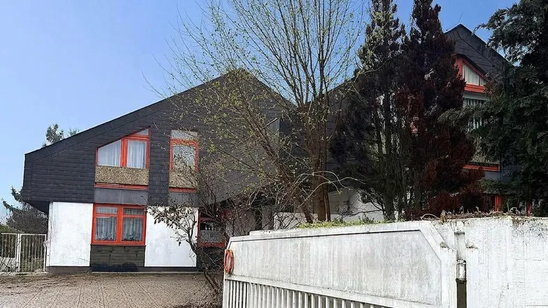 Haus mit markantem Dach, weißen Wänden und orangefarbenen Fenstern, umgeben von Bäumen und einem Zaun.