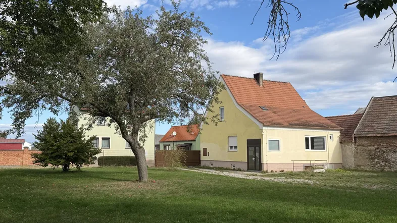 Gelbes Einfamilienhaus mit rotem Ziegeldach und großem Gartenbereich unter blauem Himmel.