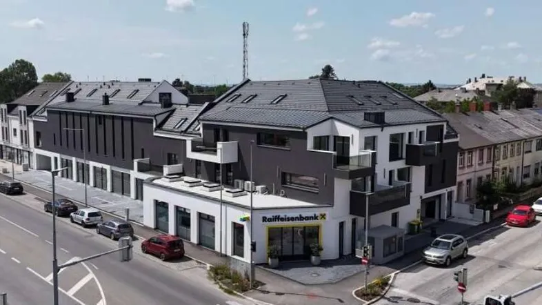 Moderne Wohn- und Geschäftsanlage mit Raiffeisenbank-Filiale an einer belebten Straße, umgeben von weiteren Gebäuden.