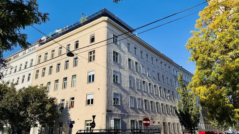 Mehrstöckiges Wohngebäude mit klassischer Fassade und Bäumen im Vordergrund an einem sonnigen Tag.