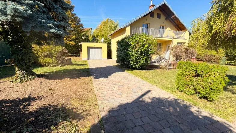 Gelbes Einfamilienhaus mit gepflasterter Auffahrt, Vorgarten und separater Garage unter blauem Himmel.