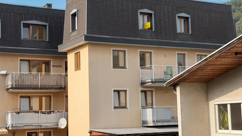 Mehrfamilienhaus mit mehreren Balkonen, Mansarddach und Bergblick im Hintergrund.