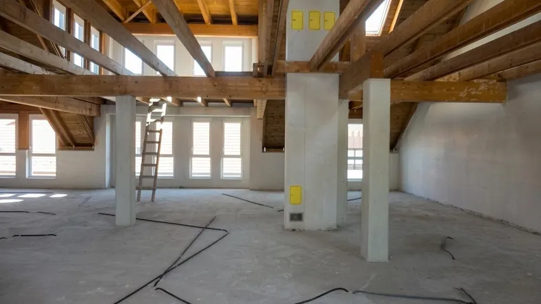 Geräumiger Dachboden im Rohbau mit sichtbaren Holzbalken, Betonboden und vielen Fenstern, die viel Tageslicht hereinlassen.