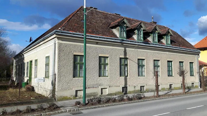 Altes Wohnhaus mit Putzfassade, grünen Fensterrahmen und Ziegeldach an einer Straße gelegen.