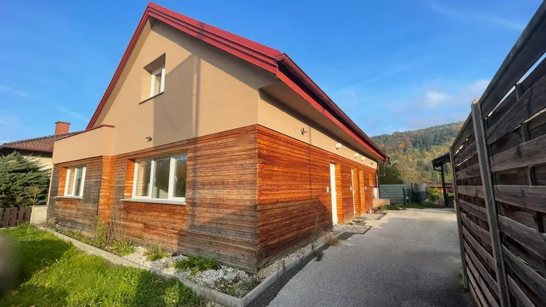 Seitenansicht des Gebäudes mit Holzfassade, rotem Dach und grüner Umgebung.