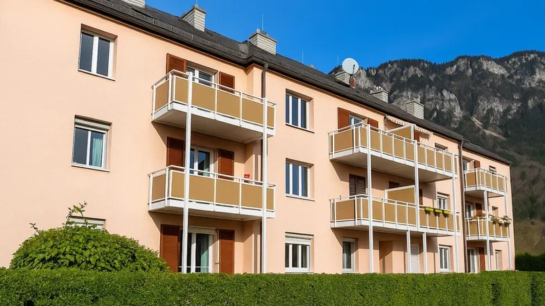 Mehrfamilienhaus mit mehreren Balkonen und grüner Hecke im Vordergrund, umgeben von Bergen.