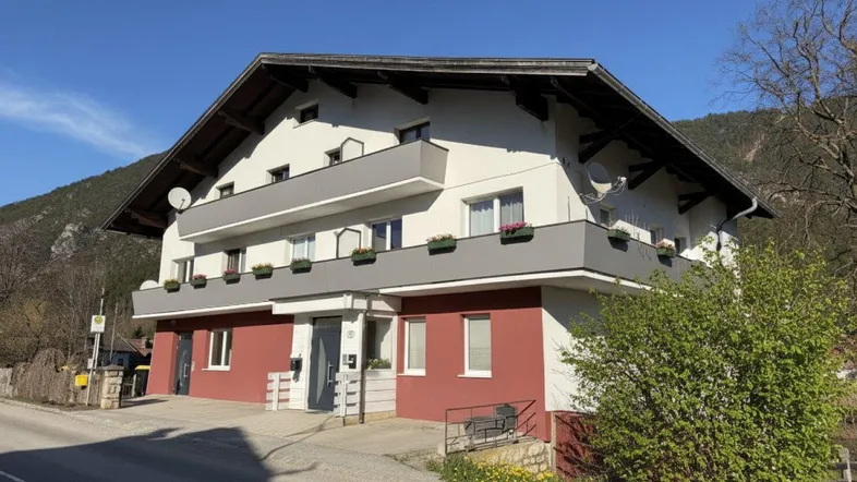 Ansicht des Mehrfamilienhauses von der Straße mit Balkonen und Bergblick unter blauem Himmel.