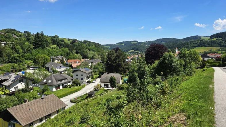 Panoramablick über eine grüne Wohnsiedlung in einer hügeligen Landschaft unter blauem Himmel.