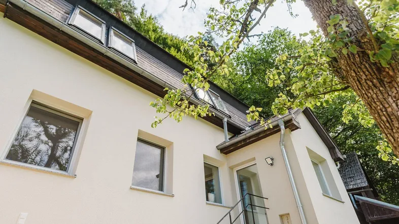 Modernes Haus mit Dachgauben und weißer Fassade, umgeben von viel Grün.
