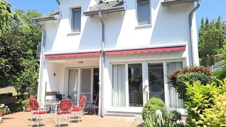 Weiße Hausfassade mit großer, gepflasterter Terrasse, roter Markise und Gartenmöbeln.
