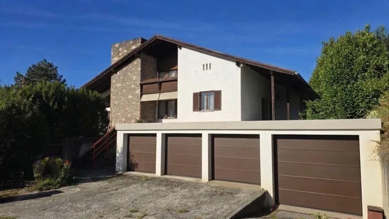 Traditionelles Haus mit Satteldach und integrierter Garage, umgeben von viel Grün.