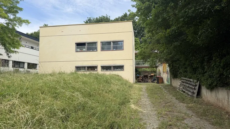 Gelbes Gebäude mit mehreren Fenstern und einer überwucherten Zufahrt, umgeben von Bäumen und Sträuchern.