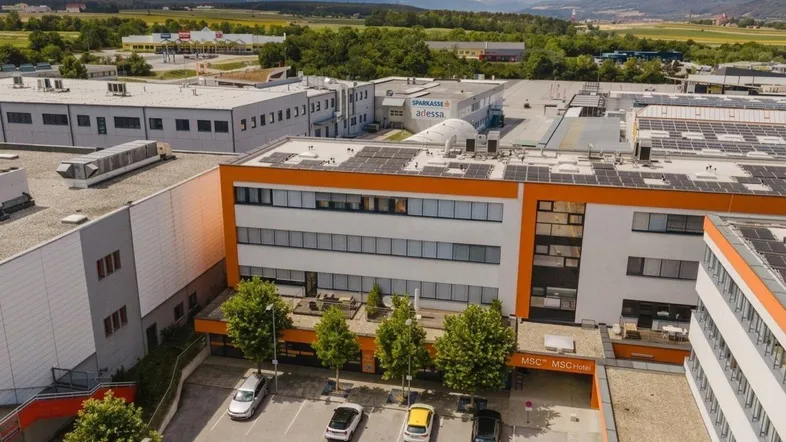 Luftaufnahme eines modernen Geschäftsgebäudes mit Solaranlagen, Parkplätzen und Bergkulisse im Hintergrund.