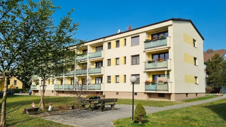 Mehrfamilienhaus mit gelber Fassade, Balkonen und gepflegter Grünanlage unter blauem Himmel.