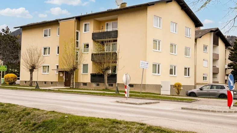 Mehrfamilienhaus mit mehreren Balkonen an einer Straße gelegen, umgeben von Bäumen.