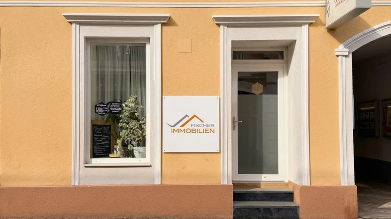 Gelbe Fassade eines Geschäftsgebäudes mit Schaufenster und Eingangstür auf einem gepflasterten Gehweg.