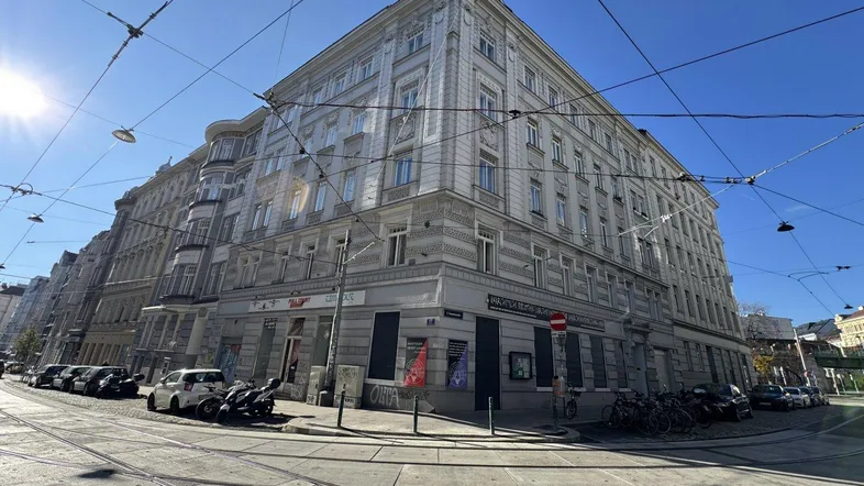Historisches Eckgebäude in städtischer Umgebung mit Straßenbahnschienen und sonnigem Wetter.