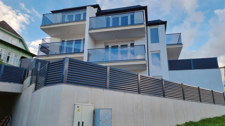 Modernes Mehrfamilienhaus mit mehreren Balkonen und Glasgeländern in einer Hanglage.