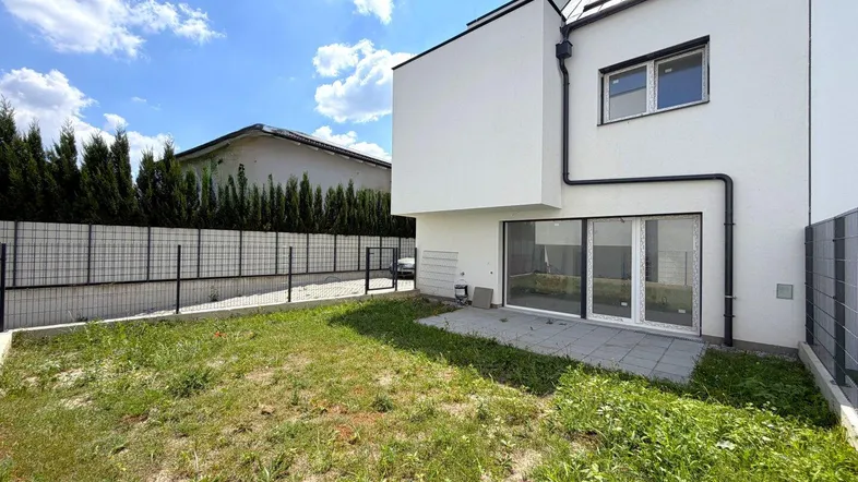 Hinterhof mit Rasenfläche und kleiner gepflasterter Terrasse, umgeben von einem Zaun.