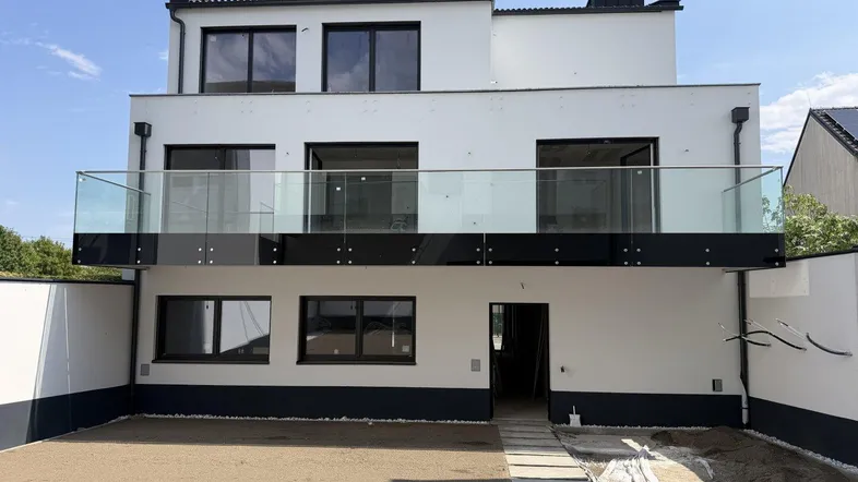 Modernes Mehrfamilienhaus mit weißer Fassade, großen Fenstern und einem Balkon mit Glasgeländer.