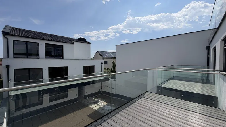 Moderne Terrasse mit Holzdeck und Glasgeländer, umgeben von Neubauten unter blauem Himmel.