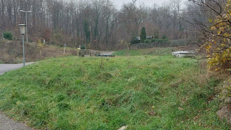 Unbebautes Grundstück mit Grasbewuchs und Bäumen im Hintergrund, an einer ländlichen Straße gelegen.