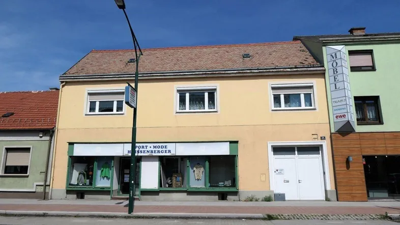 Gelbe Fassade eines Gebäudes mit Ladenlokal im Erdgeschoss und Wohnfenstern im Obergeschoss.