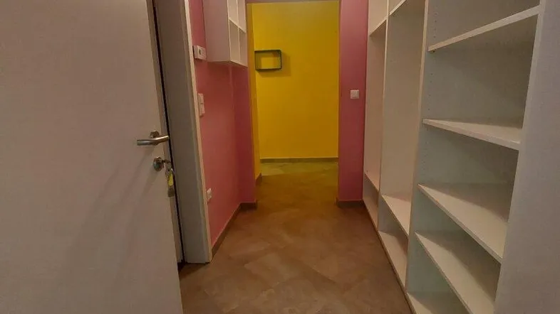 Langer Flur mit rosa Wänden und weißen Regalen, der zu einem gelben Raum führt.