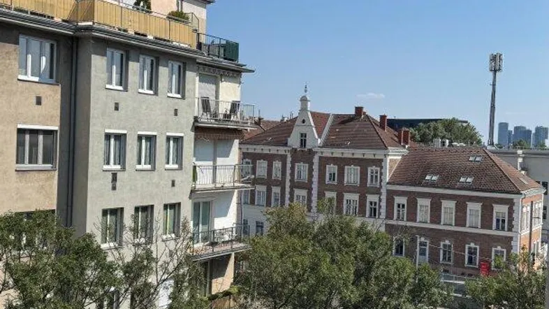 Blick vom Balkon auf die umliegenden Gebäude und grüne Bäume unter blauem Himmel.