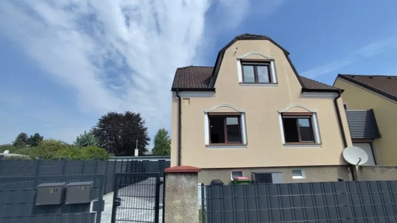 Hausfassade mit Mansarddach und Eingangsbereich, umgeben von einem modernen Zaun.
