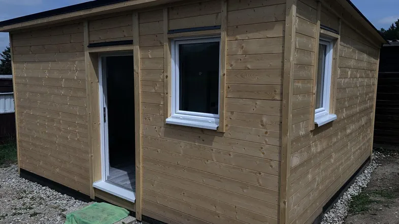 Außenansicht einer kleinen Holzhütte mit Holzwänden und Fenstern unter blauem Himmel.