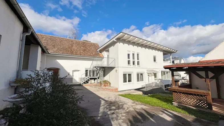 Weißes Wohngebäude mit Außentreppe und Zufahrt unter blauem Himmel, umgeben von Grünflächen.