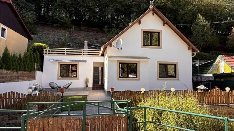 Weißes Einfamilienhaus mit Satteldach, umzäuntem Vorgarten und Blick auf bewaldeten Hang.