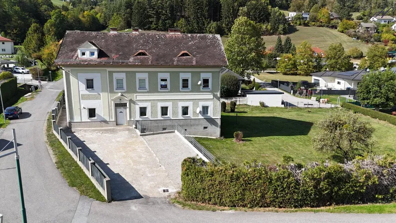 Luftaufnahme eines traditionellen Hauses mit großem Grundstück, umgeben von grüner Landschaft und weiteren Häusern.