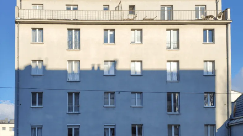 Graue Fassade eines Mehrfamilienhauses mit vielen Fenstern und einem Dachbalkon unter blauem Himmel.