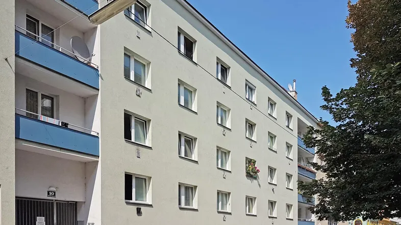 Ansicht eines Mehrfamilienhauses mit hellgrüner Fassade und blauen Balkonen an einer Straße.