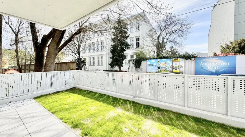 Heller Gartenbereich mit Terrasse und moderner Zaunanlage vor einem Gebäude.