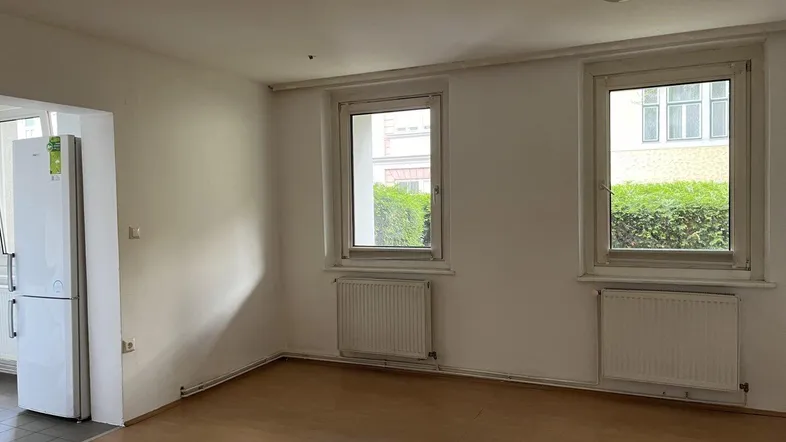 Heller Wohnraum mit Holzfußboden, zwei Fenstern und Blick auf die grüne Umgebung.