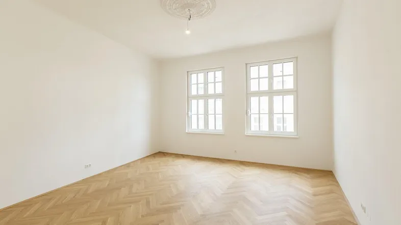 Helles, leeres Zimmer mit Fischgrätparkettboden, weißen Wänden und zwei großen Fenstern.